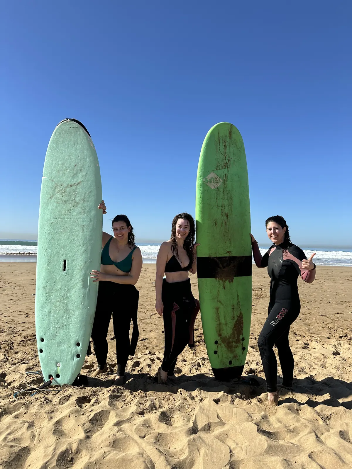 Three surfers in wetsuits standing with their soft-top boards on the beach
