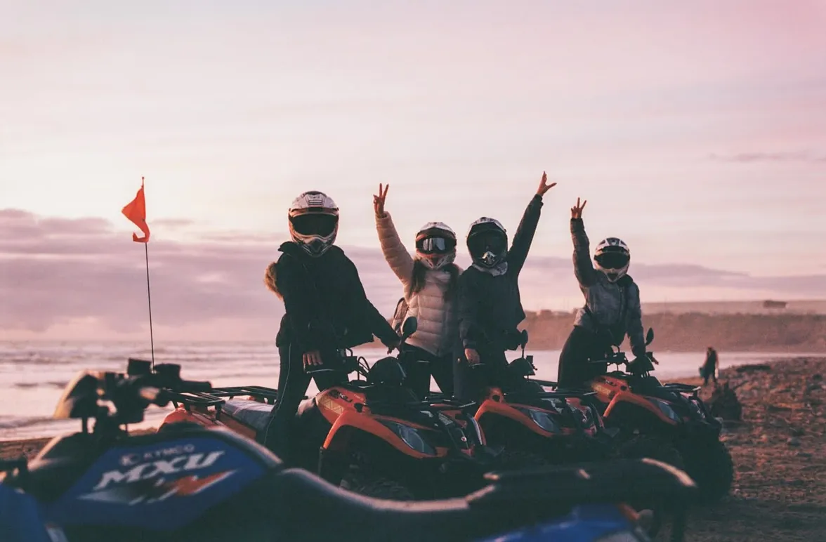 Four friends in helmets waving from their quad bikes on the beach at sunset