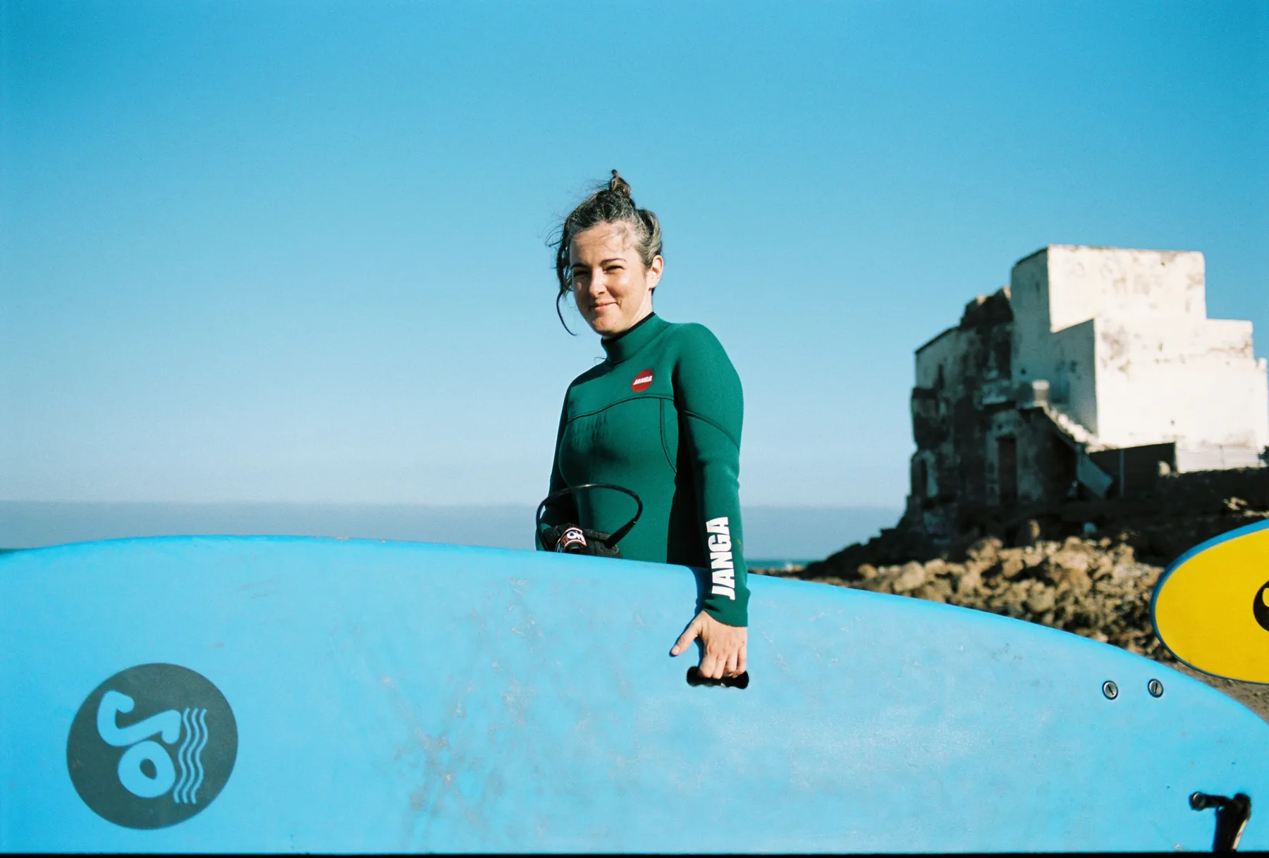 Smiling surfer in a green wetsuit holding a blue board by an old white building