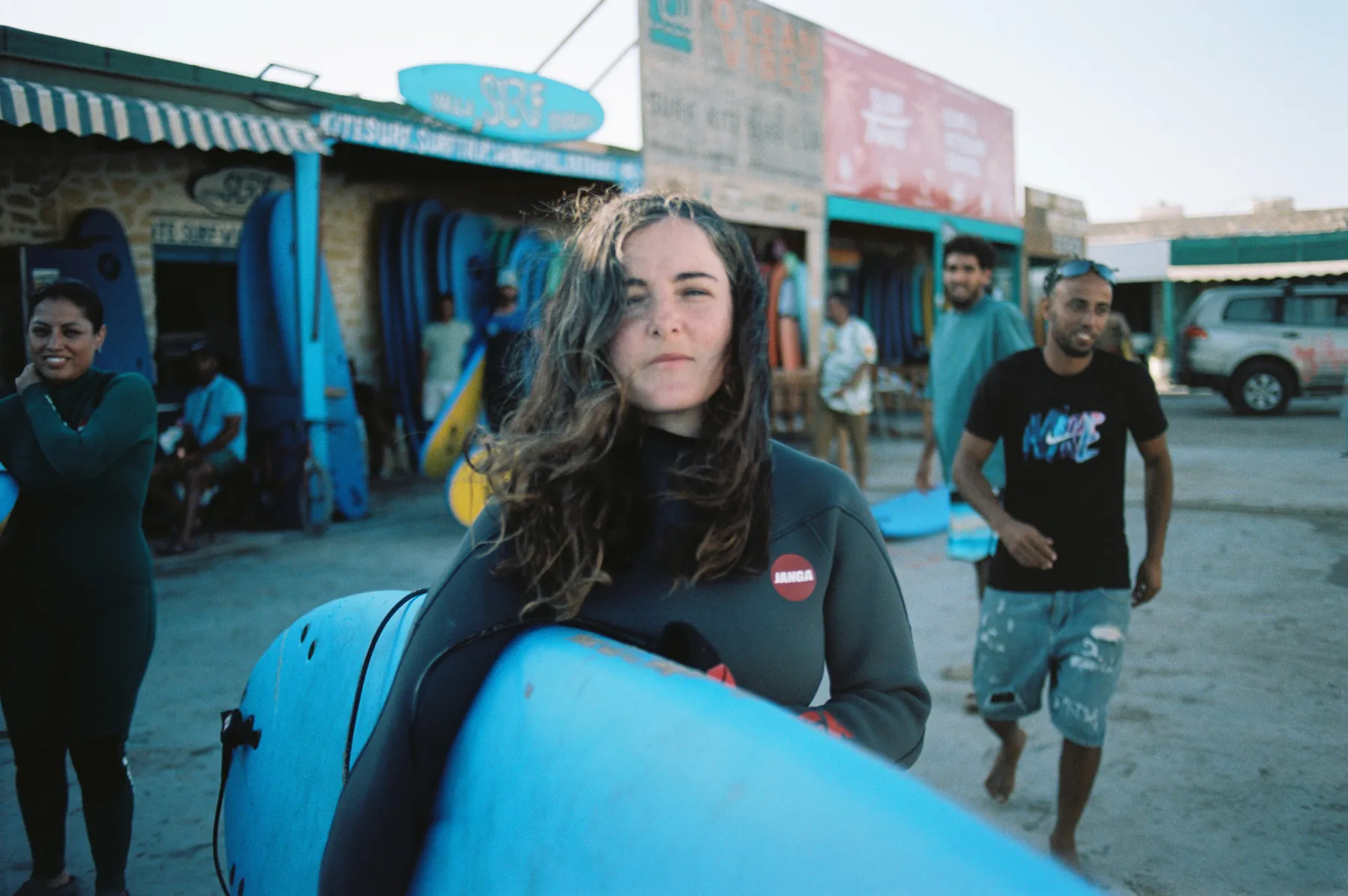Surfer with windswept hair carrying her board, friends smiling behind her