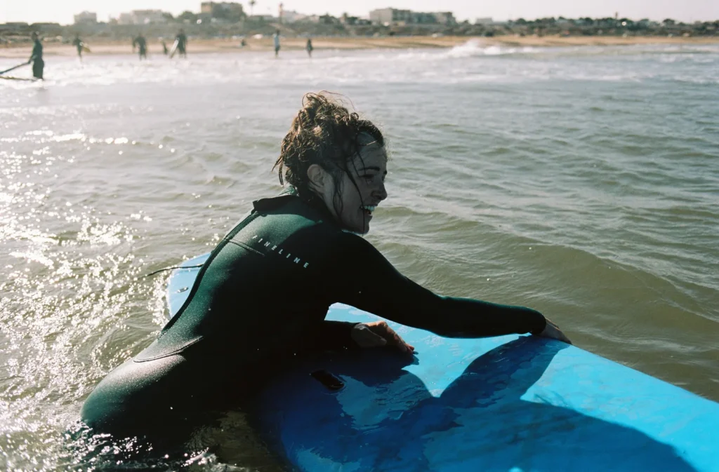 Smiling surfer in a dark wetsuit pushing her blue soft-top through the shallow water