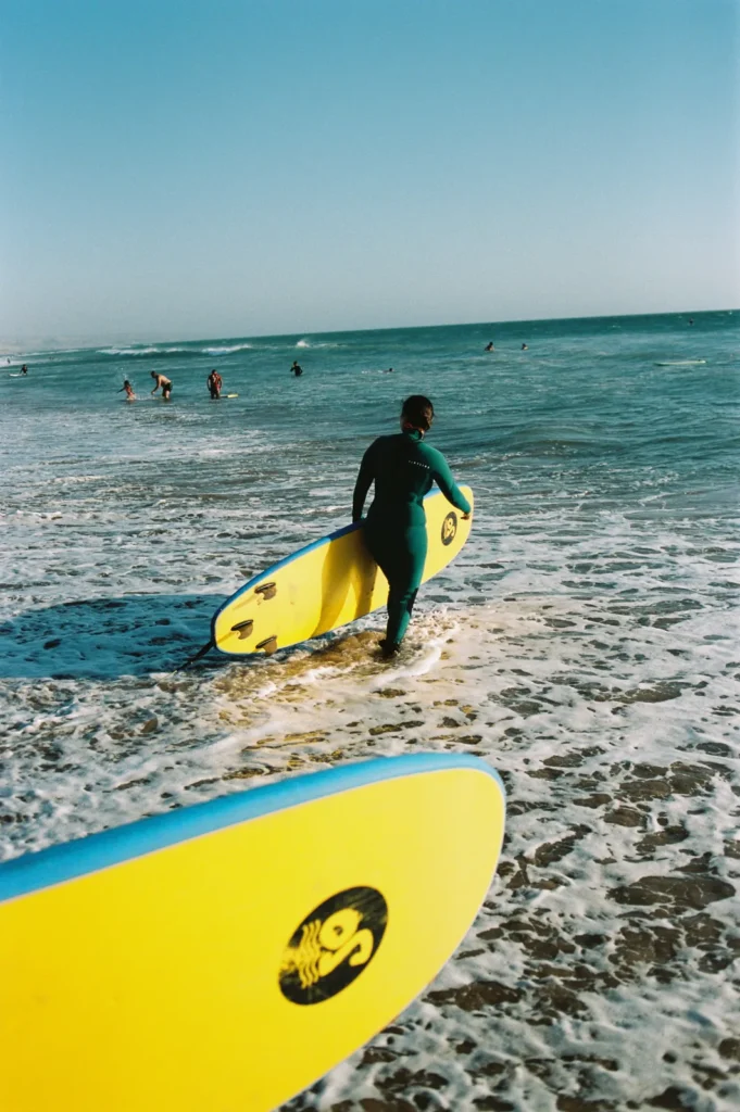 Surfer in a green wetsuit walking into the sea with a yellow soft-top, another board in the foreground