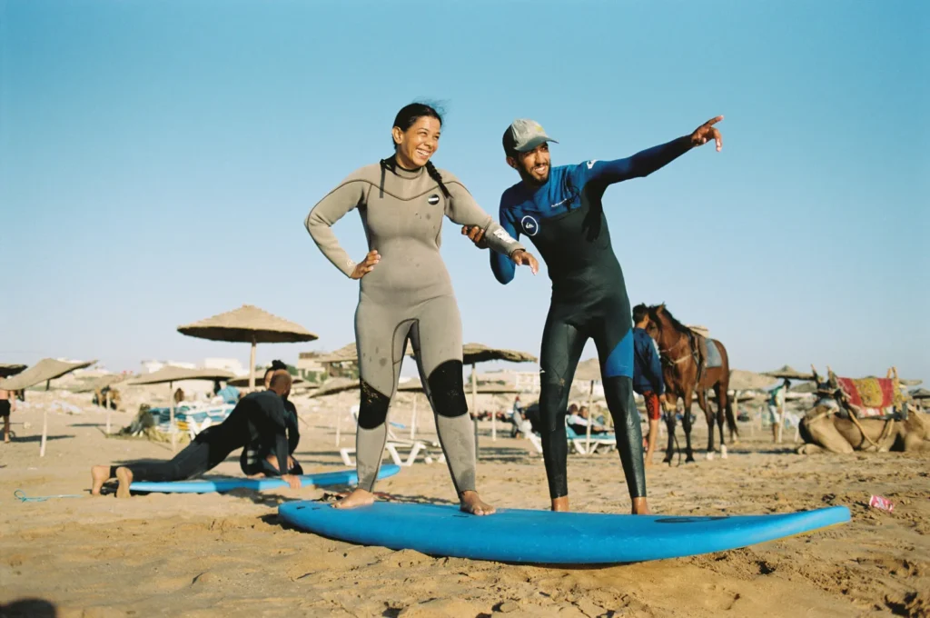Surf coach showing a laughing student how to stand on a board on the sand, umbrellas and a horse behind