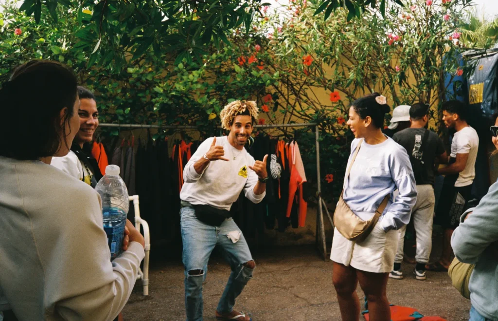 Grinning surf instructor throwing a double shaka at guests in a garden, wetsuits hanging on a rack behind